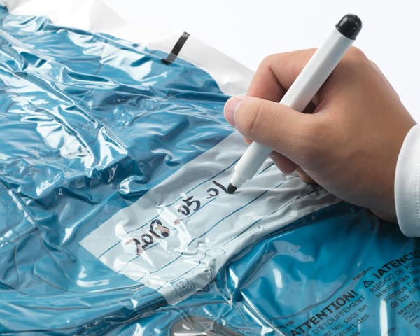 A hand writing the date 2018-05-01 on a SPANTAD vacuum seal bag filled with blue textiles.