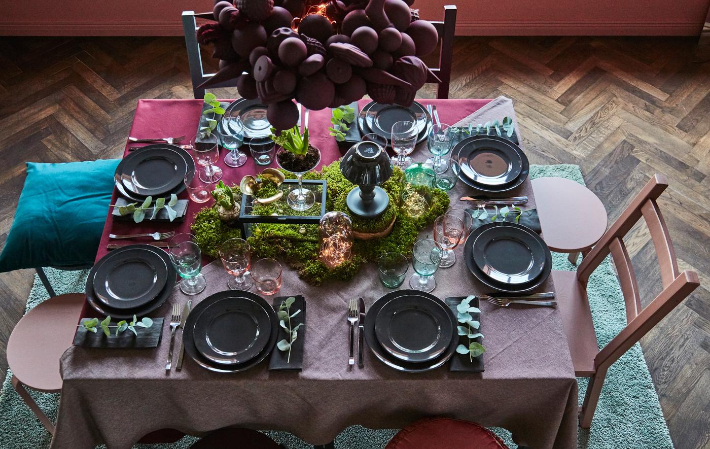 A holiday table is set using dark reds and greens, and greenery.