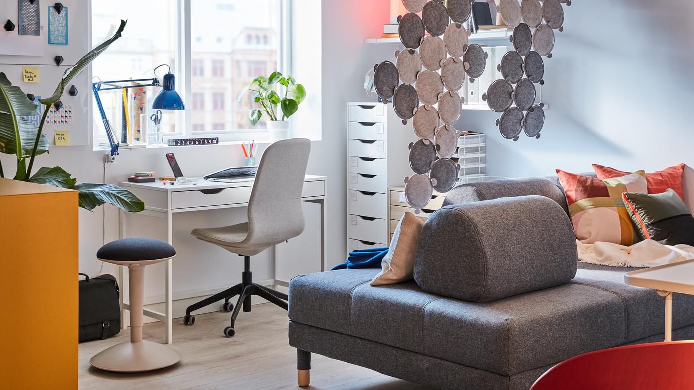 A home office in the living room with white desks and drawers, grey work chair, grey sofa and sound-absorbing panels.