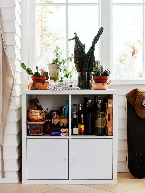 A KALLAX shelving unit by a window, organised as a small pantry with pots, dry goods, canned food, and a few plants on top.