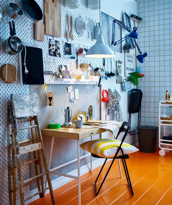 A kitchen area with perforated wall tiles and kitchen utensils hanging from hooks, with a breakfast table and folding chairs.