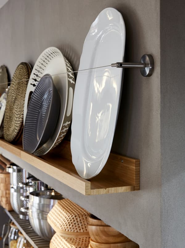 A kitchen storage wall with a wooden MÅLERÅS picture ledge displaying stoneware plates, trays and other kitchen items.