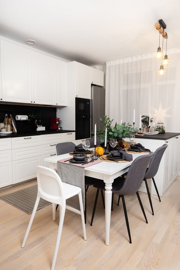 A kitchen with a dining table, mixed-style chairs, and a decorated table setting; white kitchen cabinets and pendant lights are visible in the background.