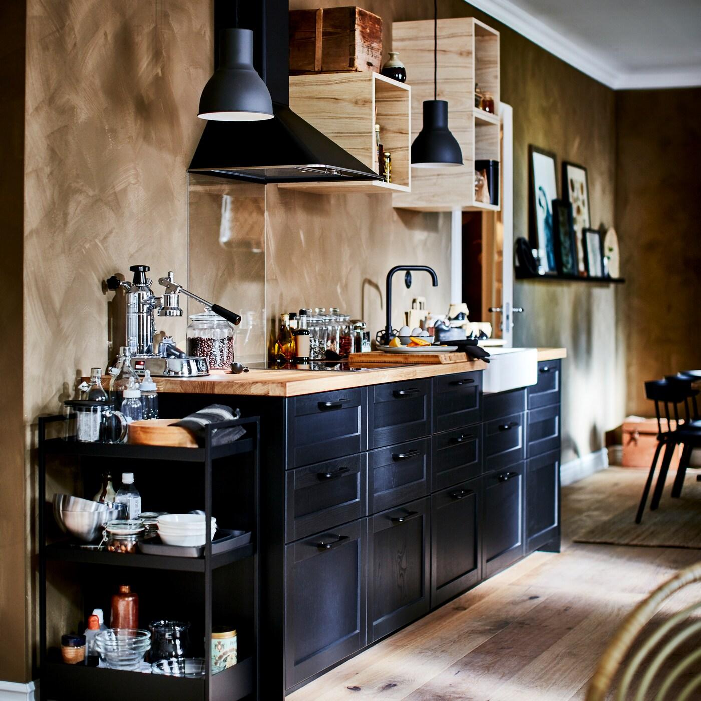 A kitchen with black drawer fronts, a wooden worktop, a black trolley, open wall cabinets and two dark grey pendant lamps.