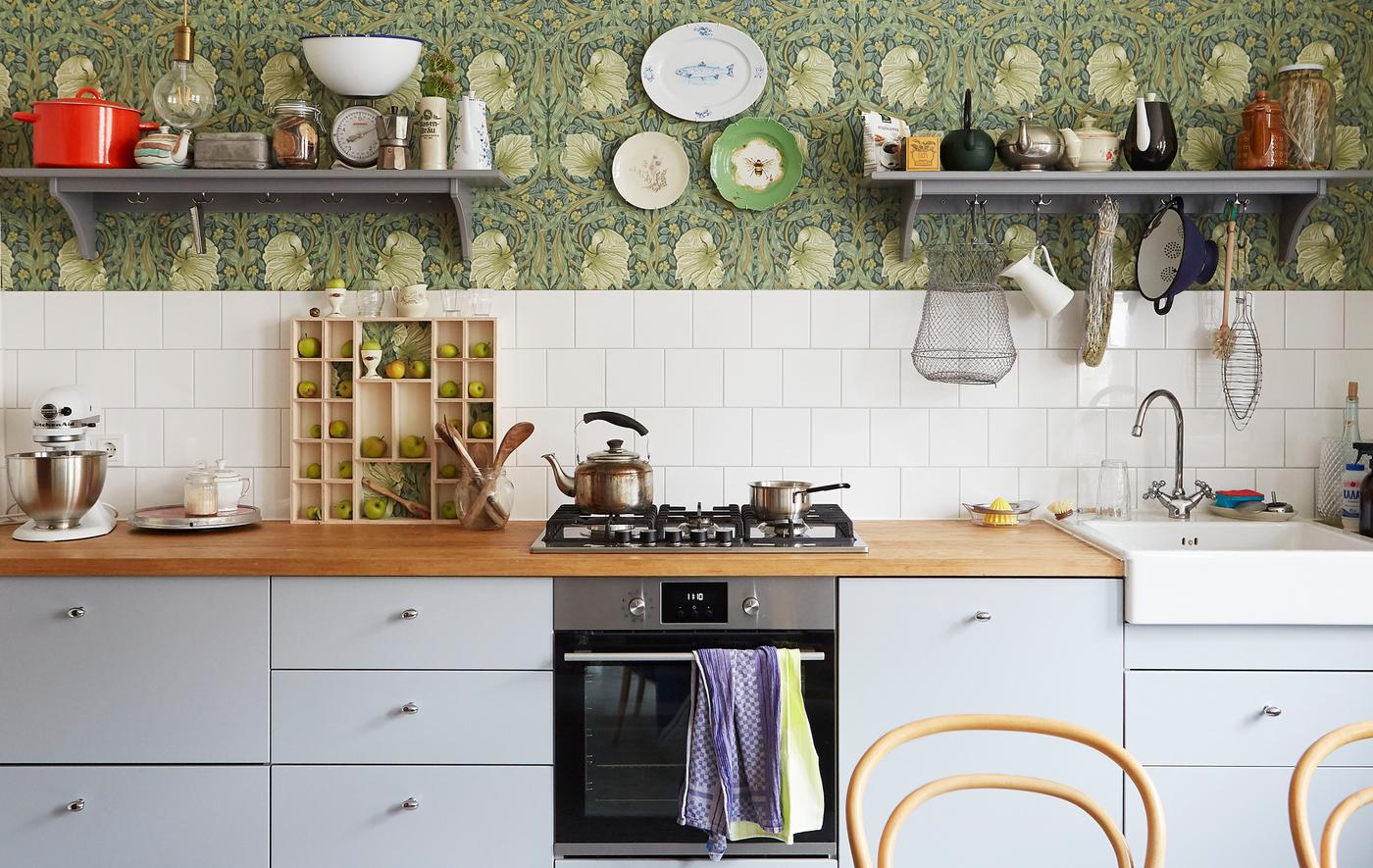 A kitchen with grey cabinets and built-in cooker, wooden worktops, and shelves on patterned floral wallpaper.