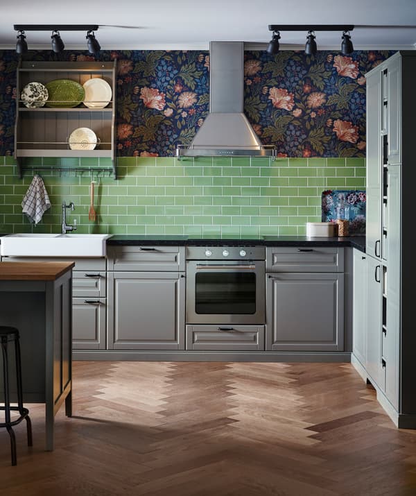 A kitchen with grey cabinets, green tiles and floral wallpaper, with a stainless steel extractor hood.