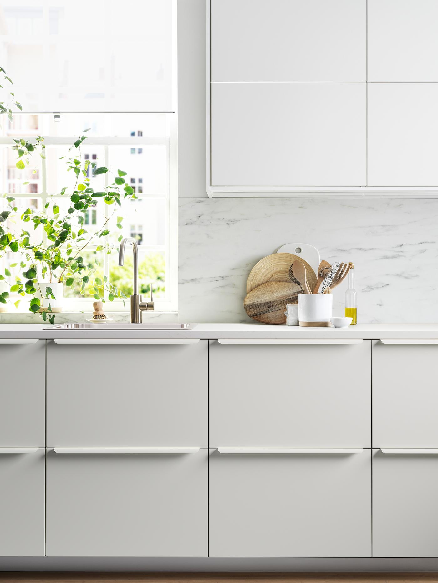 A kitchen with white cabinet fronts, a white worktop, a white marble-effect/quartz splashback and a plant next to a window.