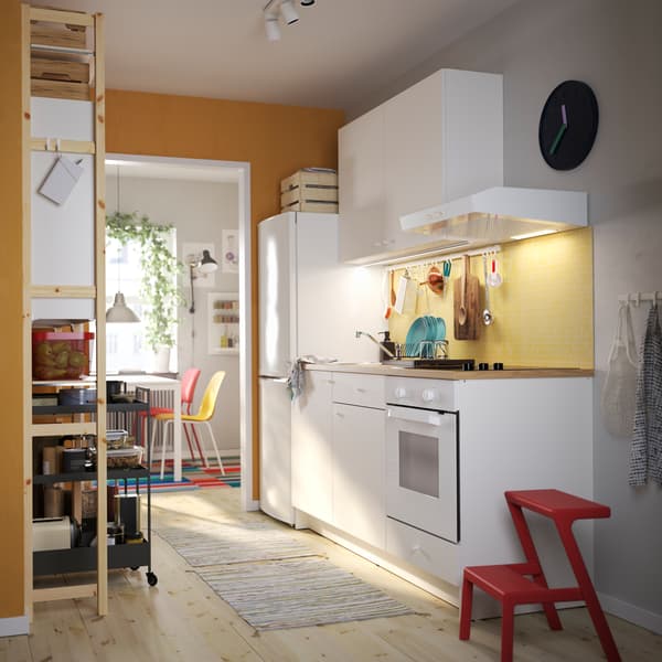A KNOXHULT kitchen in white, a red step stool, a shelving unit in pine, black trolleys and colourful kitchen chairs.
