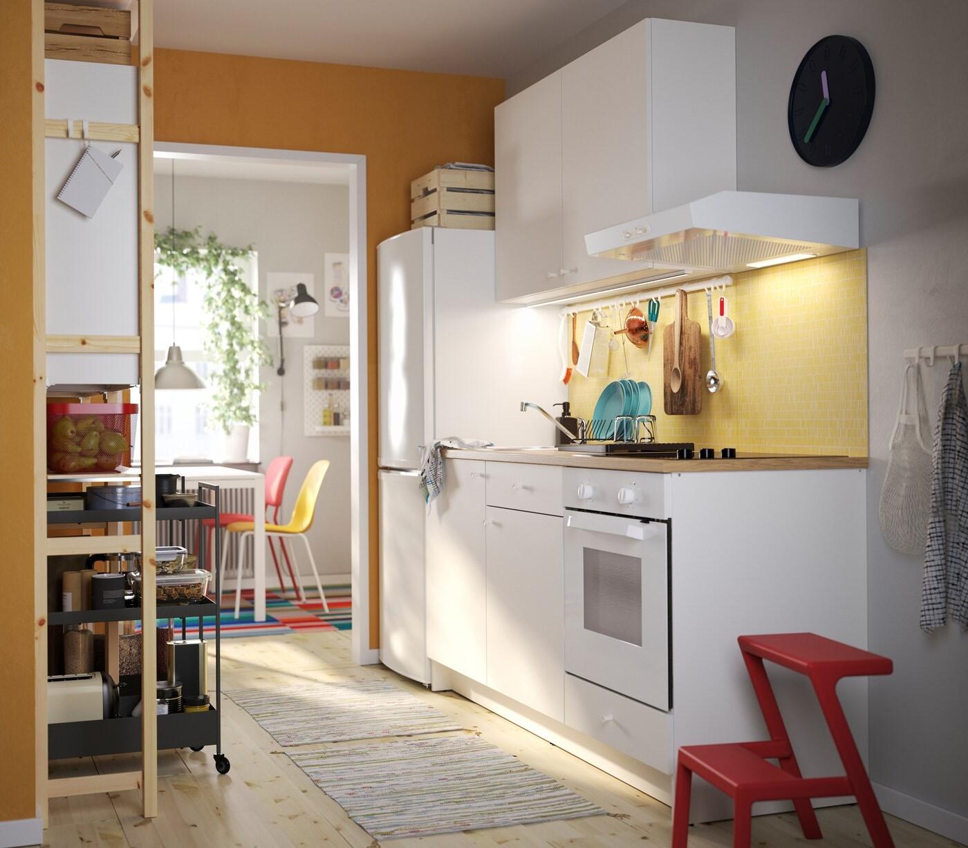 A KNOXHULT kitchen in white, a red step stool, a shelving unit in pine, black trolleys and colourful kitchen chairs.