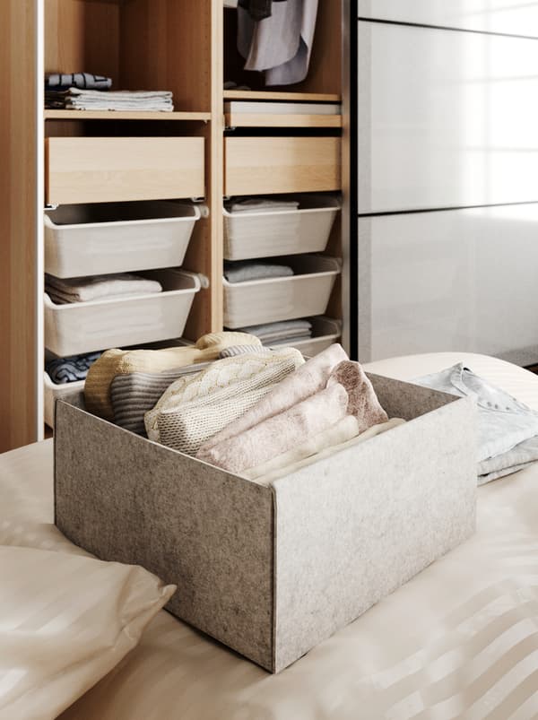 A KOMPLEMENT box on a bed, holding neatly folded garments, behind it an open wardrobe with both drawers and hanging clothes.