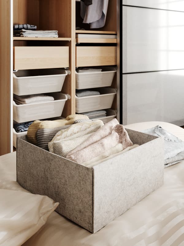 A KOMPLEMENT box on a bed, holding neatly folded garments, behind it an open wardrobe with both drawers and hanging clothes.