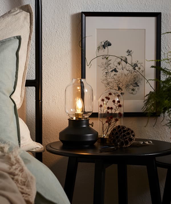 A KRAGSTA table placed bedside, decorated with a dried plant in a glass dome and a TÄRNABY lamp with dimmable LED light.