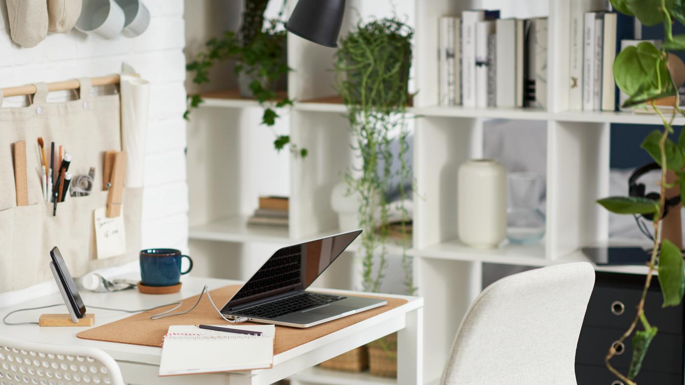 A laptop on a white table with one white and one beige chair, beside a white shelving unit with books, plants and storage.