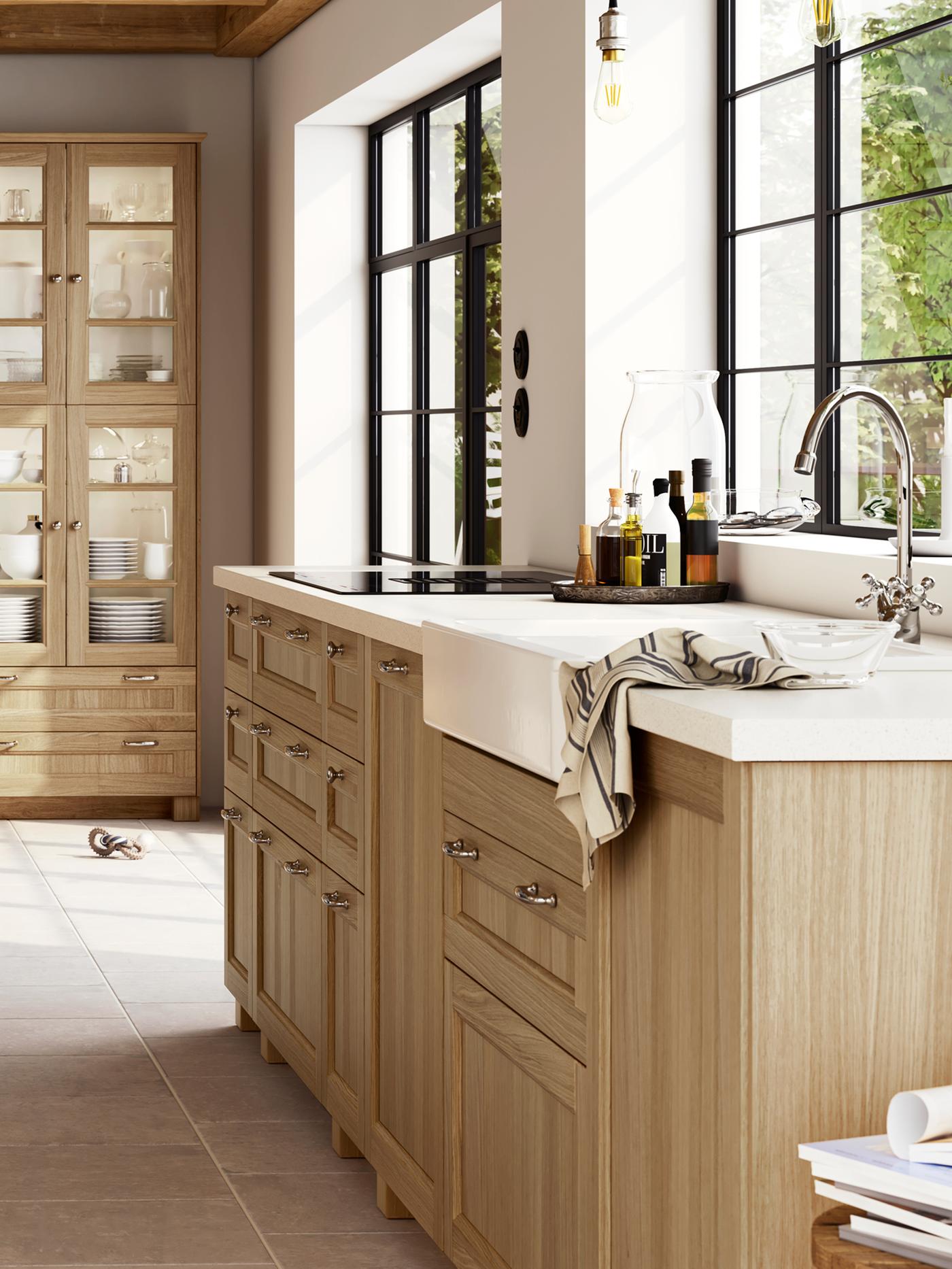 A large, bright kitchen with oak VEDHAMN fronts, including glass door cabinets filled with glassware and dinnerware.