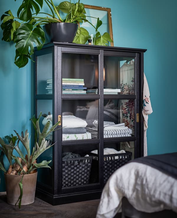 A large glass cabinet surrounded by plants is filled with books and linens.