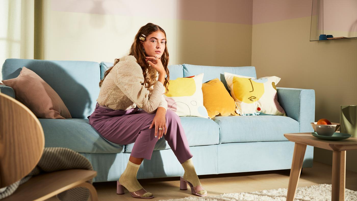 A large light-blue sofa with a variety of cushions and a young woman sitting there, in a modern, pastel living room.