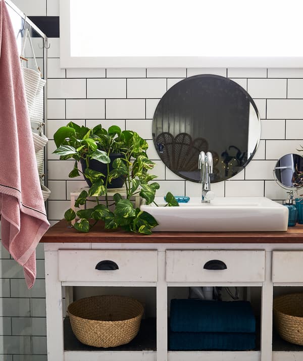 A large white sink and plant on a white vanity unit with wooden top, with a round mirror on a white tiled wall above.