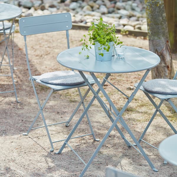 A leafy plant in a SOCKER plant pot placed on a grey SUNDSÖ table sided by grey SUNDSÖ chairs in a sunny outdoor space.