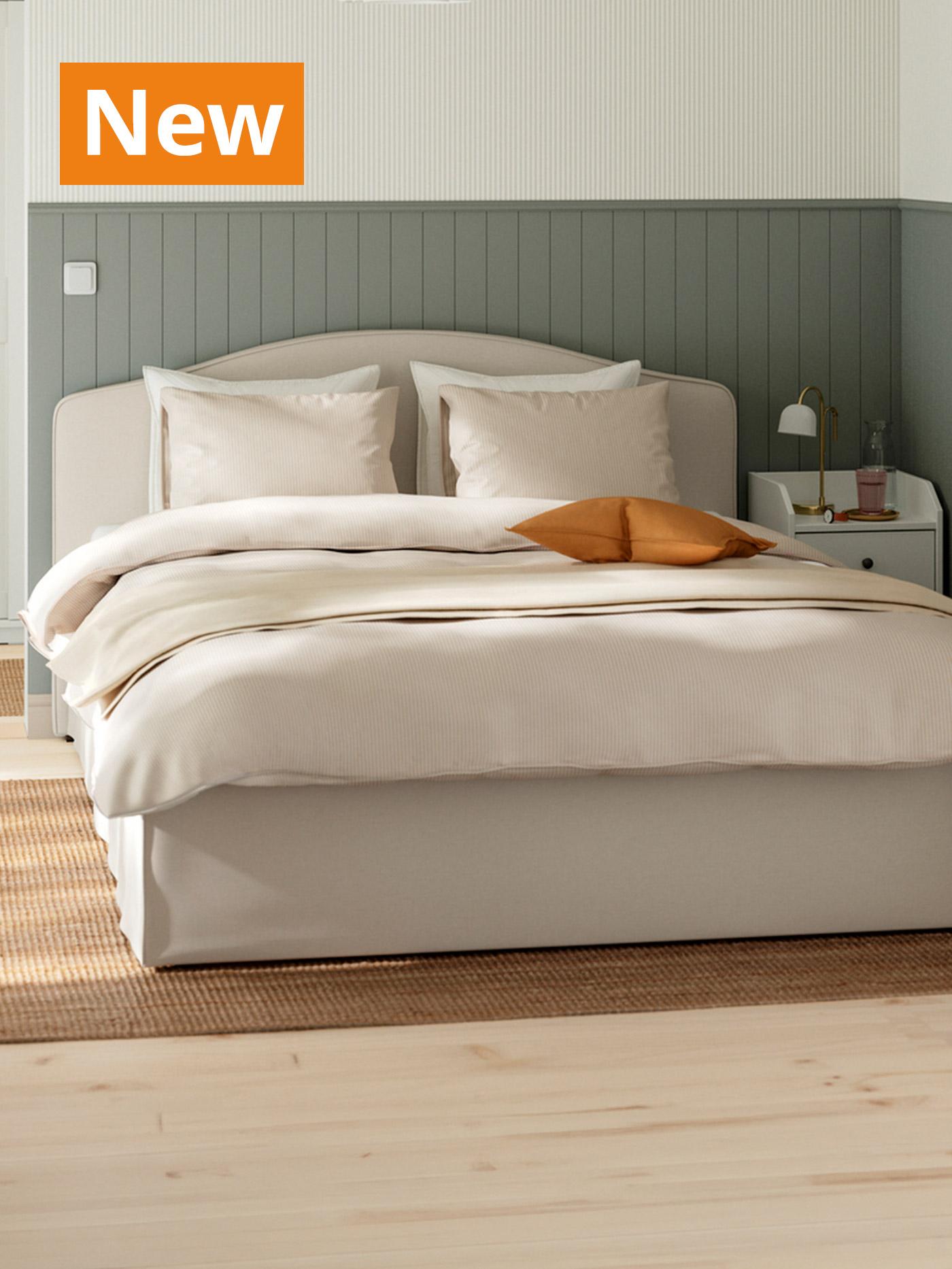 A lifestyle image of a beige/white RAMNEFJÄLL upholstered bed in a corner of the bedroom setting. Overlaying the image, orange tag that reads 'New' on the top left corner.