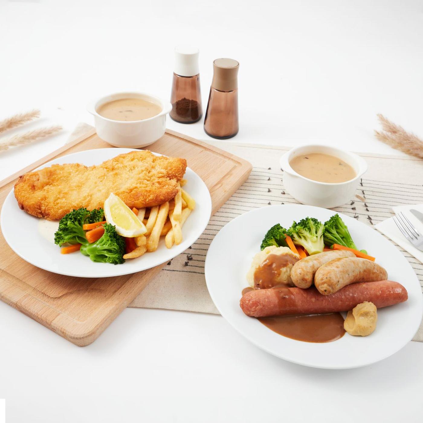 A lifestyle shot of October Feast Special happening at IKEA Singapore - Banger & mashed (2 pcs of German Pork Bratwurst & 1 pc Smoked Chicken Garlic Bratwurst with mashed potato, mixed vegetables with green soya bean served with cream sauce & mild mustard) and Pork Schnitzel with fries and mixed vegetables with green soya beans, served with a lemon wedge and aioli sauce.