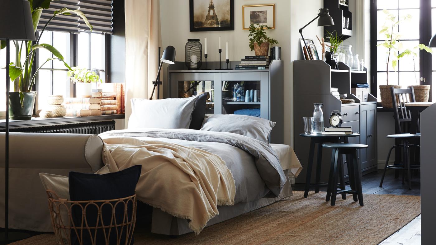 A light beige sofa-bed made as a bed with grey bedding. Behind is a grey glass-door cabinet and a jute rug lies on the floor.