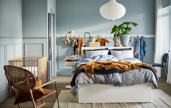 A light blue bedroom with a white bed, a white lamp, a white panel of hooks, a rattan rocking chair and a SNIGLAR baby cot.