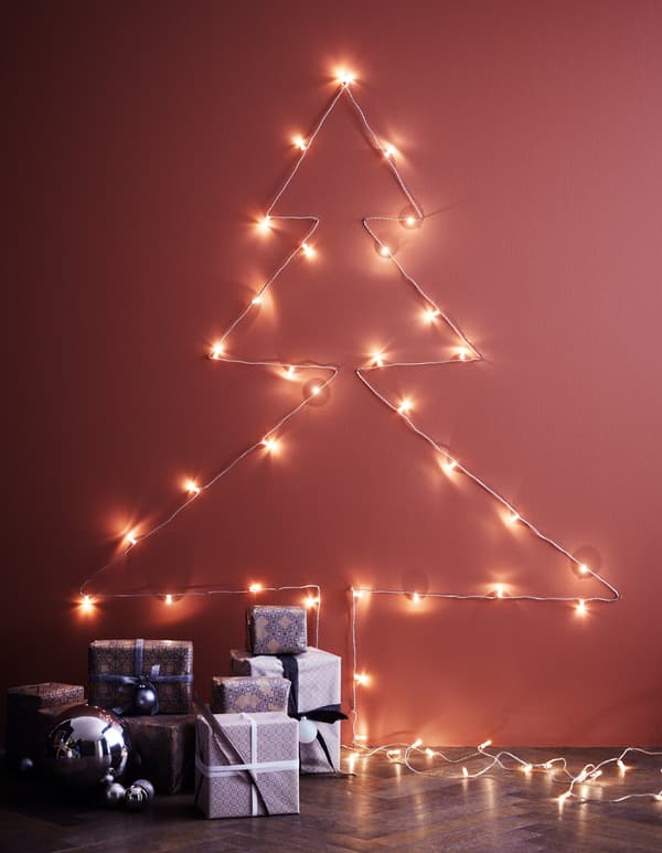A light chain trailed across a wall in the shape of a Christmas tree, using nails as support and with gifts on the floor.