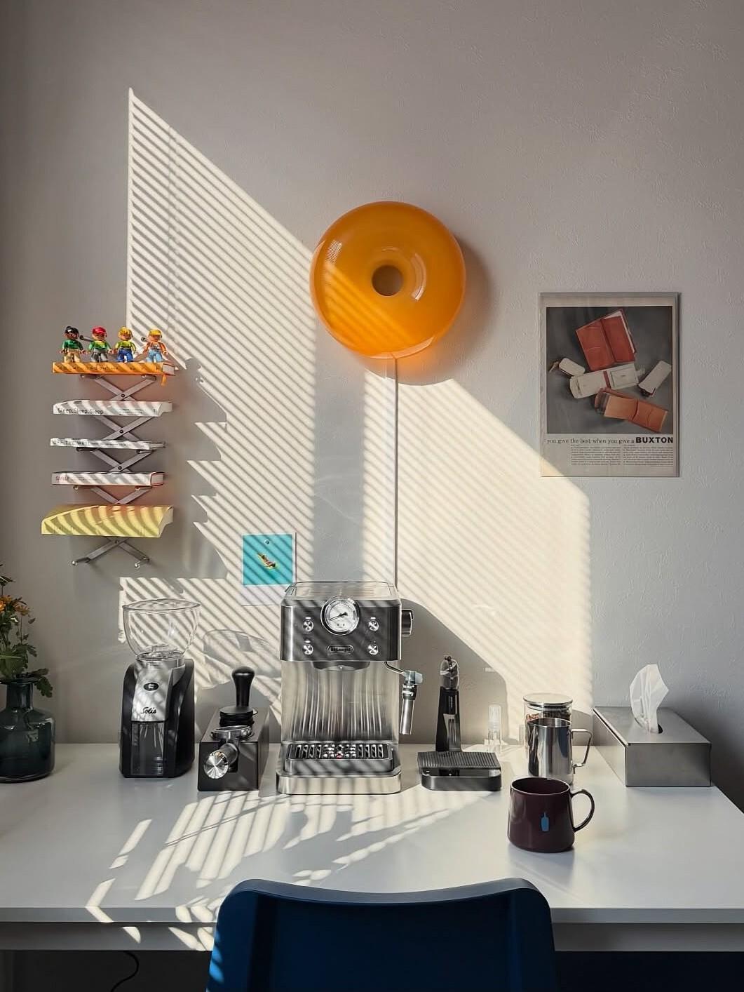 A light coffee corner with an espresso machine on a white desk, a coffee grinder and cups, a bright yellow wall lamp, decorative shelves, and sunlight casting stripes on the wall.