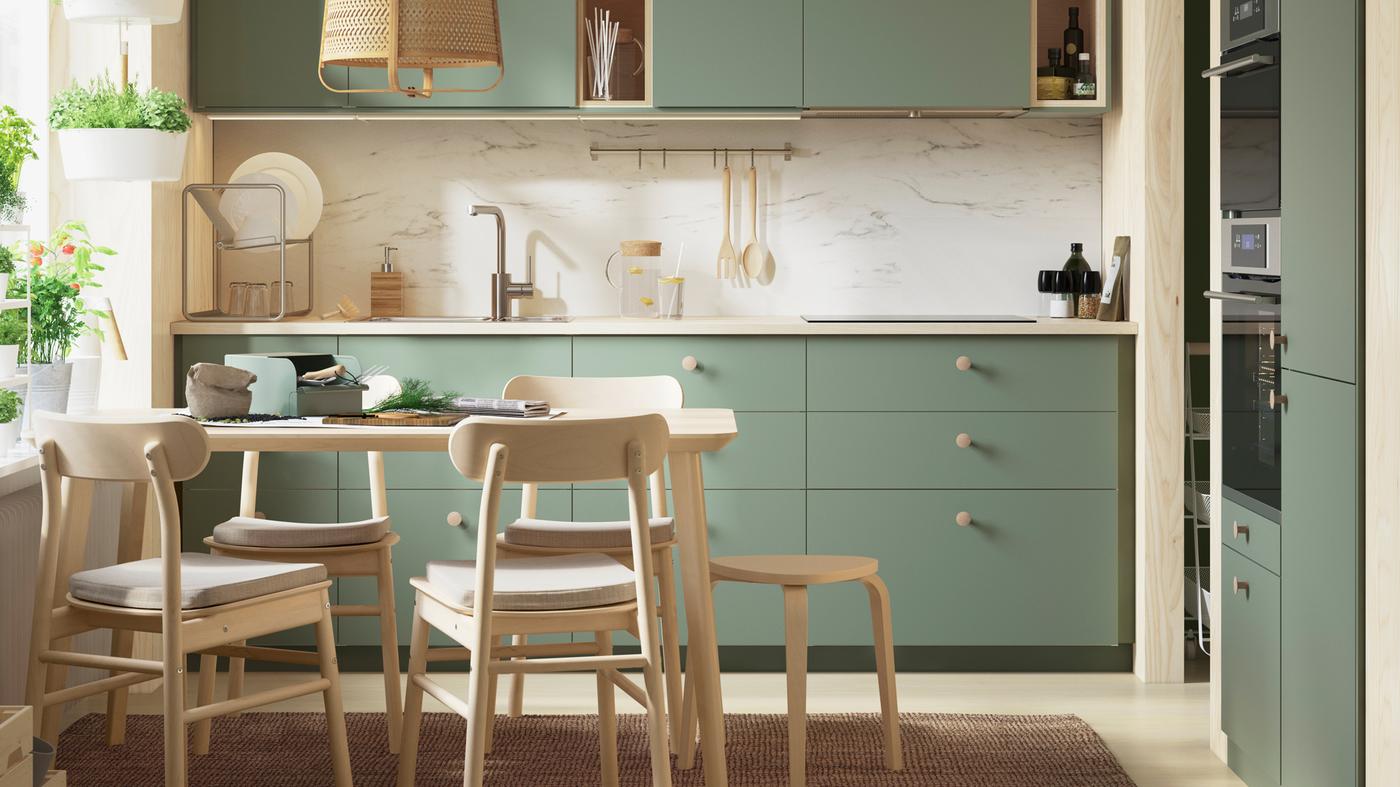 A light-drenched grey-green BODARP kitchen with herbs growing in the window, a table and four RÖNNINGE chairs in birch.