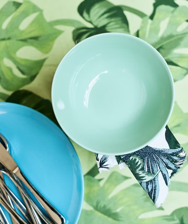 A light green bowl next to a turquoise plate with cutlery on top, on a table covered with green leaf-print fabric.