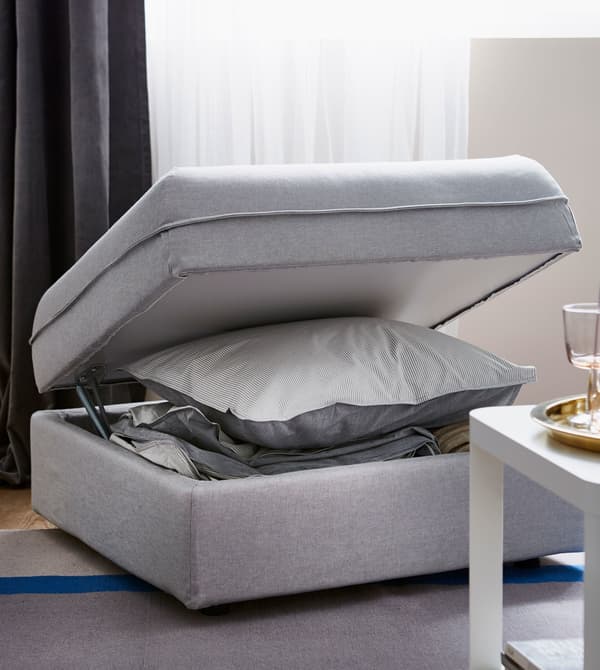 A light grey IKEA VALLENTUNA modular sofa unit, with lid opened to reveal a storage compartment for pillows and bedding.
