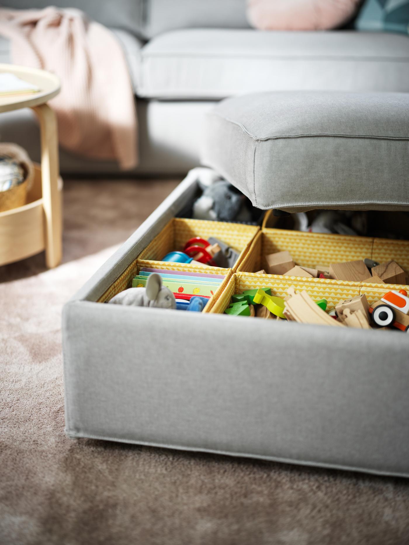 A light-grey KIVIK footstool with storage with its lid half-off, revealing toys sorted in multiple yellow UPPRYMD boxes.