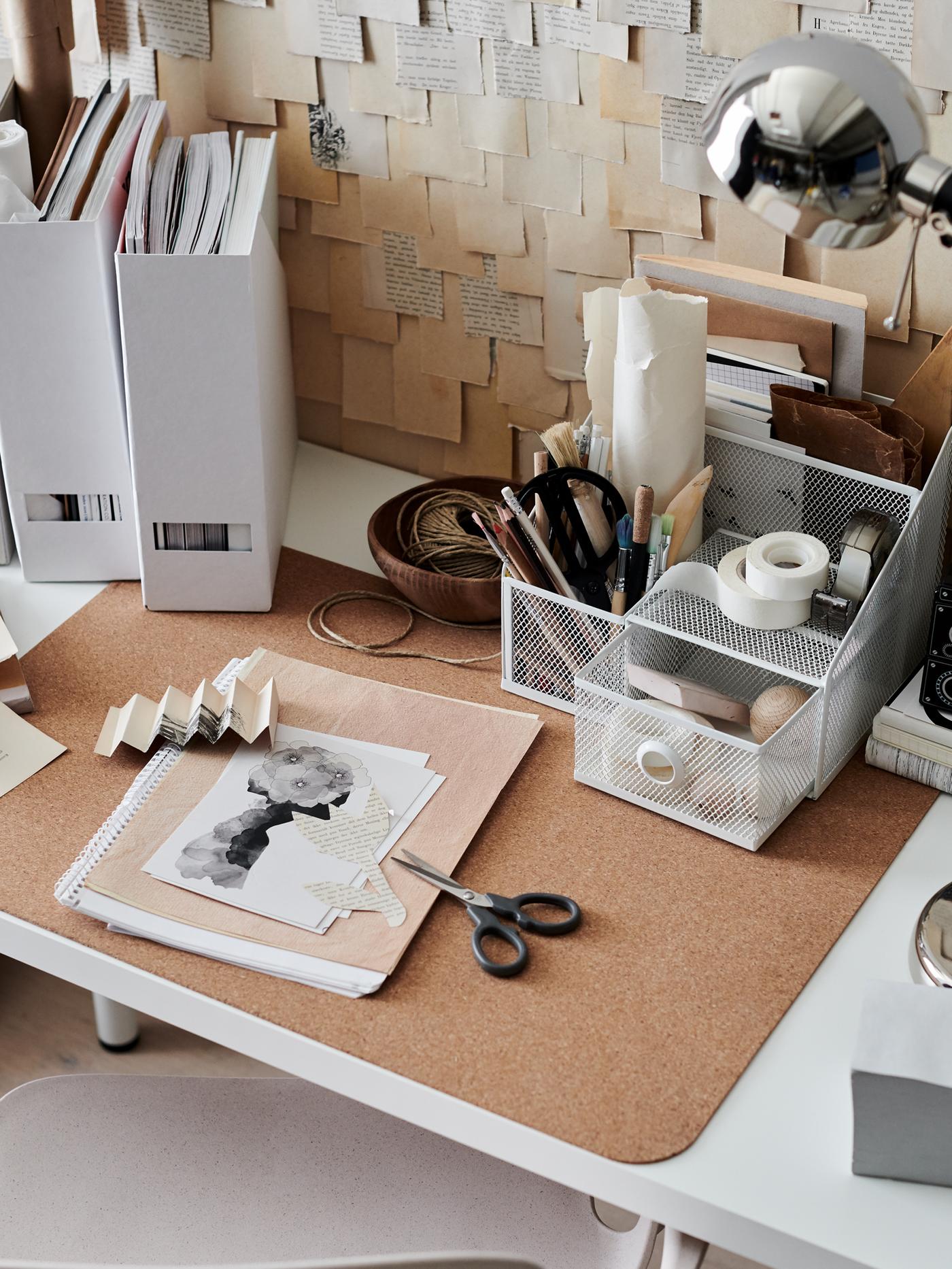 A light, somewhat cluttered workspace with a DRÖNJÖNS desk organiser, a SUSIG cork desk pad and a FORSÅ nickel-plated lamp.