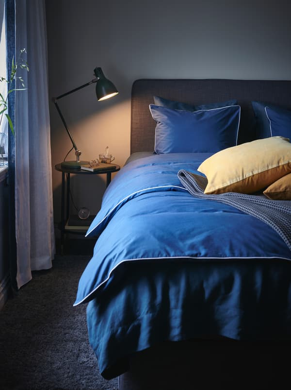 A lit reading light on a bedside table. Beside it is a bed with a blue duvet, some throw pillows and a grey bedspread.