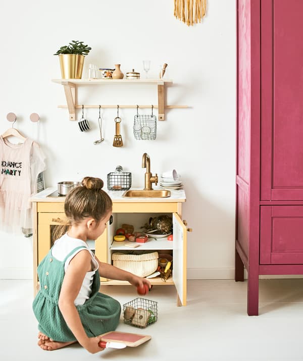 A little girl plays with a mini kitchen with accessories including toy pans, food and cooking utensils hanging from a shelf.