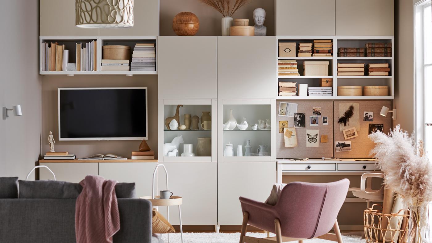 A living room with a gray-beige BESTÅ storage system with open and hidden storage, a sofa, a TV and a workstation.