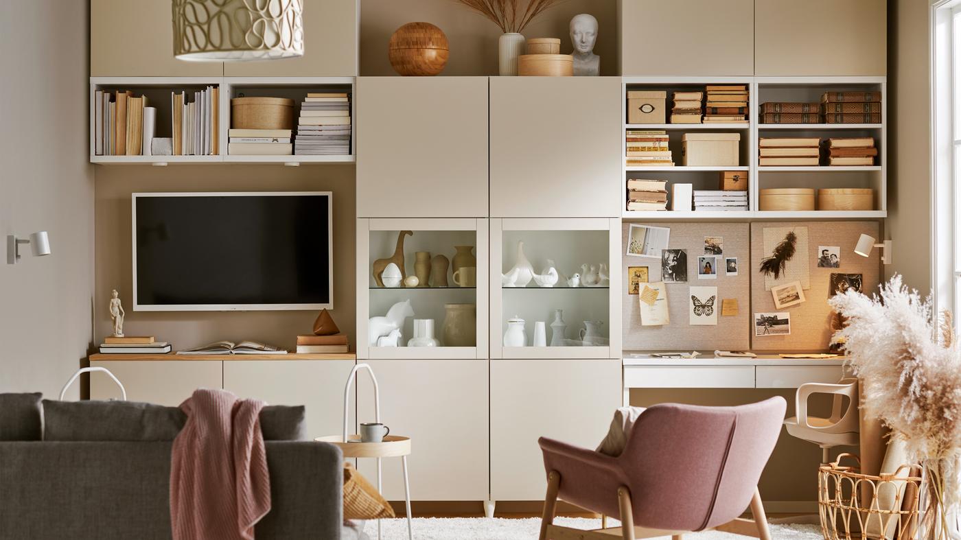 A living room with a grey-beige BESTÅ storage system giving the wall open and hidden storage, a sofa, a TV and a workstation.