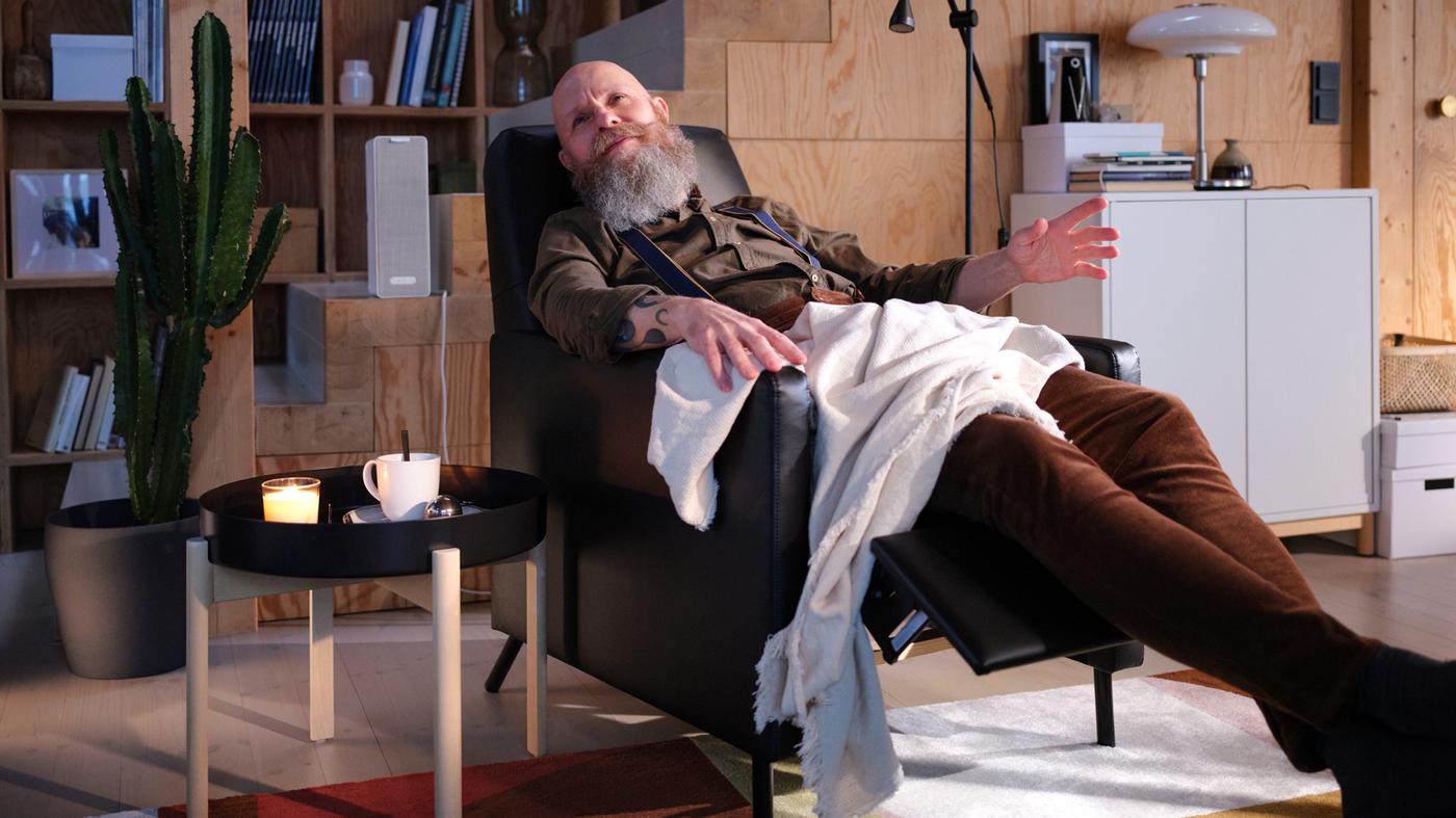 A living room with a man sitting in a black GISTAD recliner listening to music beside a coffee table with a cup of tea.