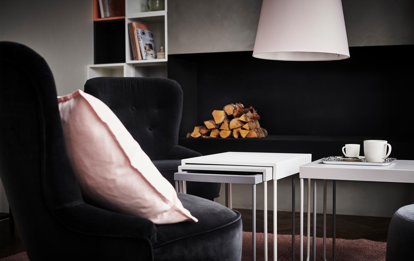 A living room with black armchairs and two sets of coffee table nests, with logs for a fire in the background.