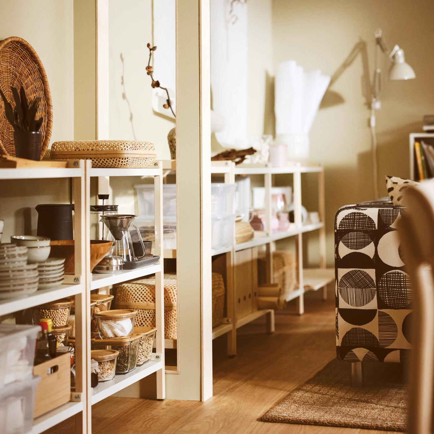 A living room with EKENABBEN open shelving units containing GLADELIG bowls, a HÖGMODIG coffee maker, and boxes with lids.