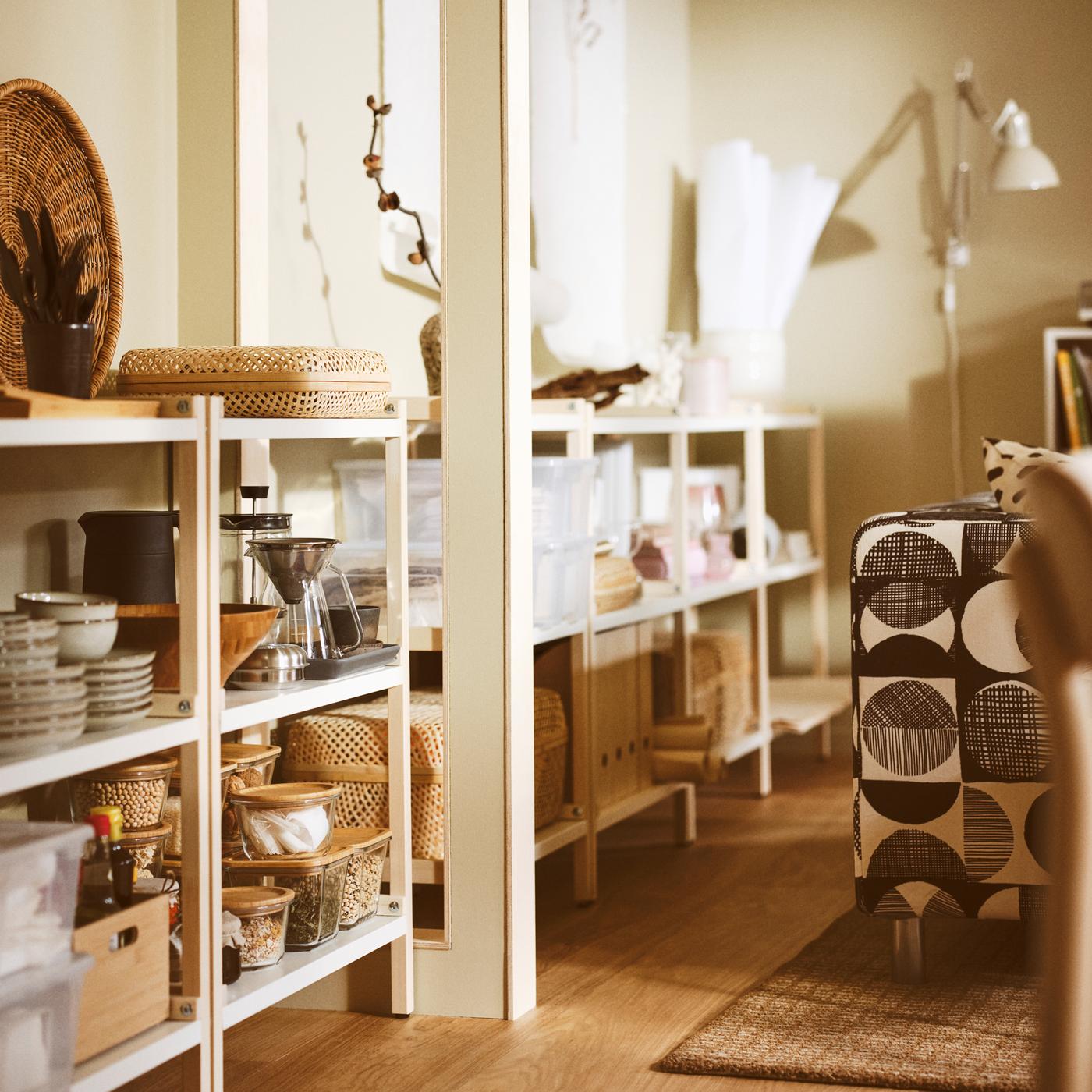 A living room with EKENABBEN open shelving units containing GLADELIG bowls, a HÖGMODIG coffee maker and boxes with lids.