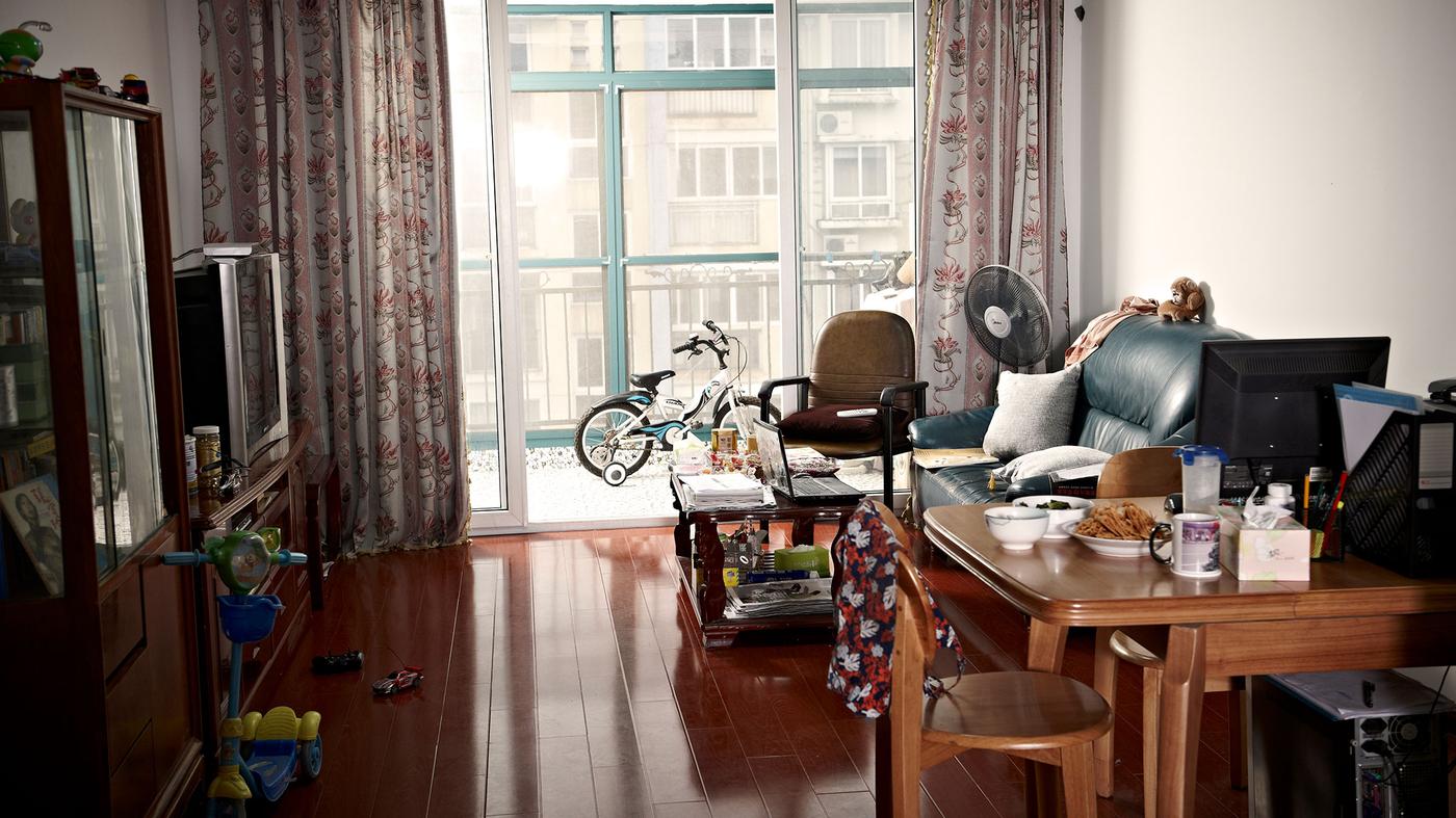 A local living room setting with a black sofa and a coffee table facing the television. The dining table is packed with local dishes and a computer for working at home. Toys are on the floor and a scooter is placed by the cupboard.