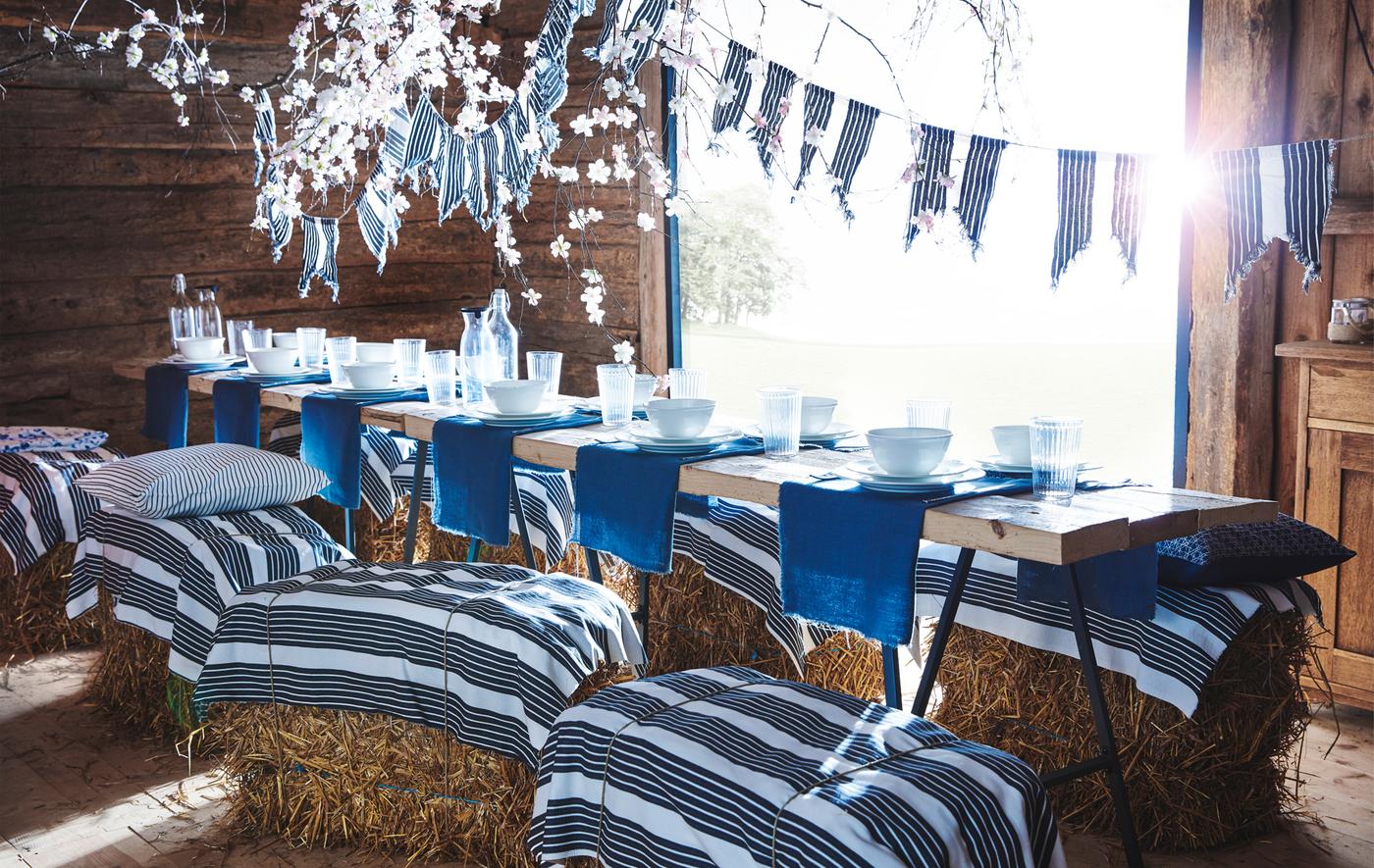 A long table in a wooden barn with blue and white tableware, and striped fabric used as bunting and tied on hay bales used as seats.