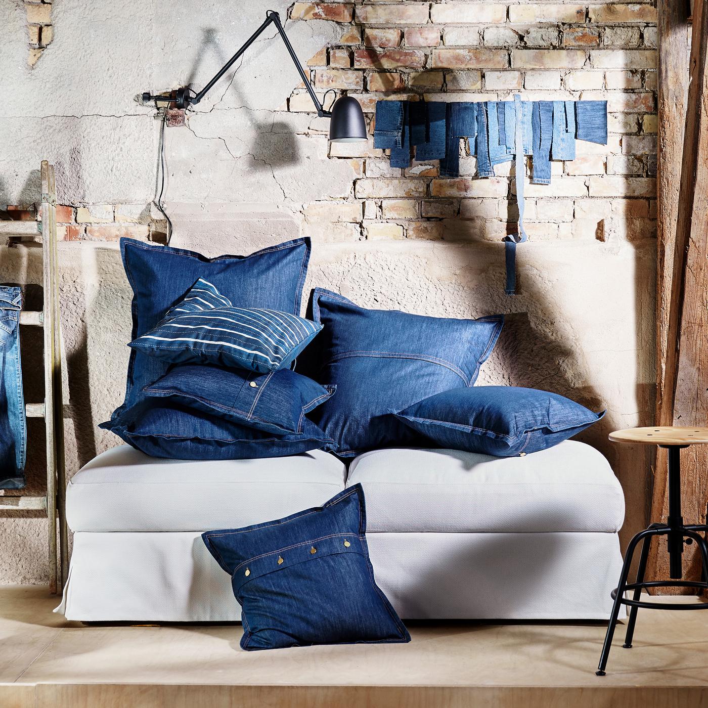 A lot of cushions in blue SISSIL cushion covers lie on top of two footstools against a wall in a run-down building.