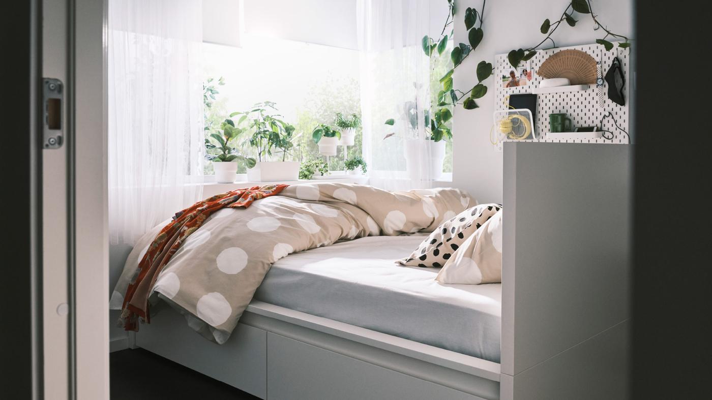 A MALM bed facing a large sunny window with lush plants on the sill. A SKÅDIS pegboard is by the bed holding various items.