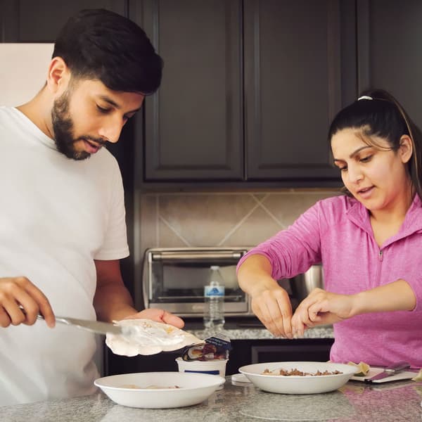 A man and a woman with one plate each are preparing breakfast in a kitchen with a stone worktop and grey cabinets.