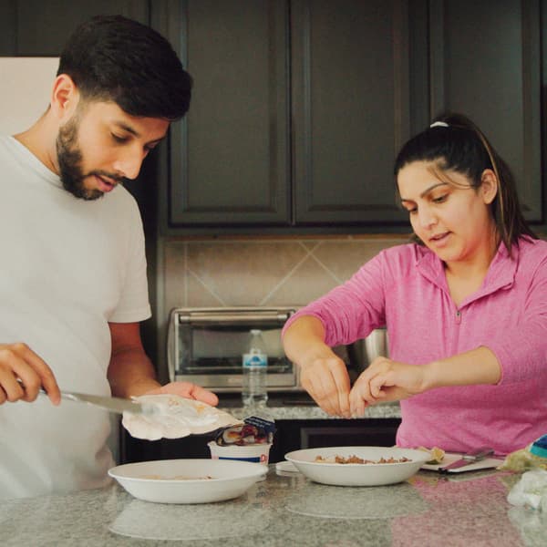 A man and a woman with one plate each are preparing breakfast in a kitchen with a stone worktop and grey cabinets.