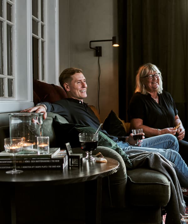 A man and woman on a green sofa with cushions and throws, drinks in glassware, tealights and books are on the coffee table.