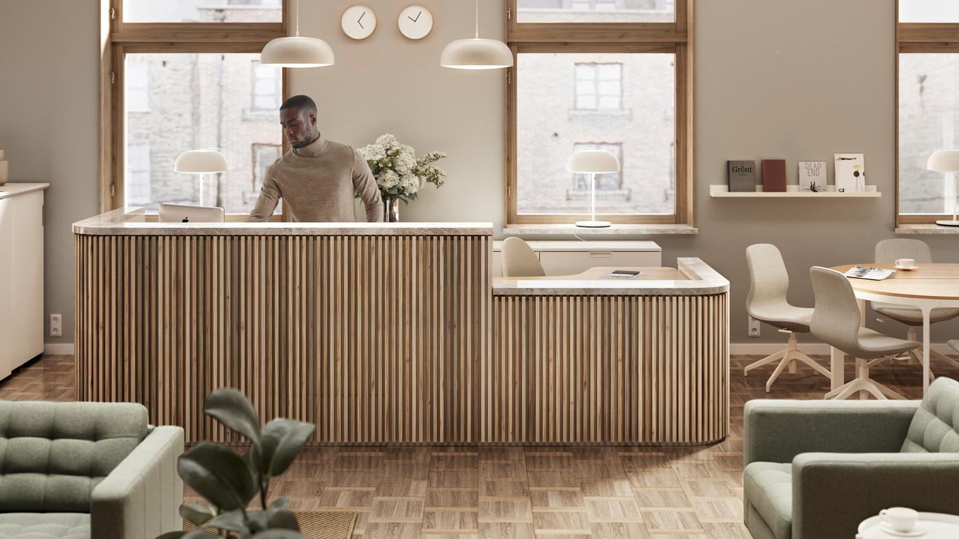 A man at a light wood reception desk in front of two windows. Two pendant lamps are above and clocks are on the wall.