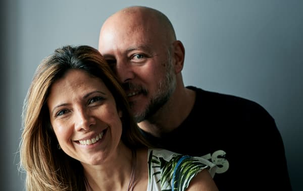 A man in a black T-shirt with a shaved head hugs a woman with brown highlighted hair.
